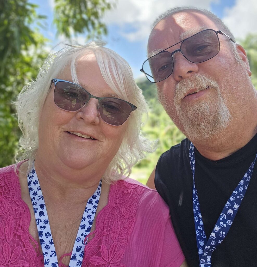 Brenda Moore, owner of Just Cruising Along next to her husband Jamie. Brenda is wearing a pink top with a blue and white lanyard. Both are wearing sunglasses and looking relaxed at their vacation by the beach